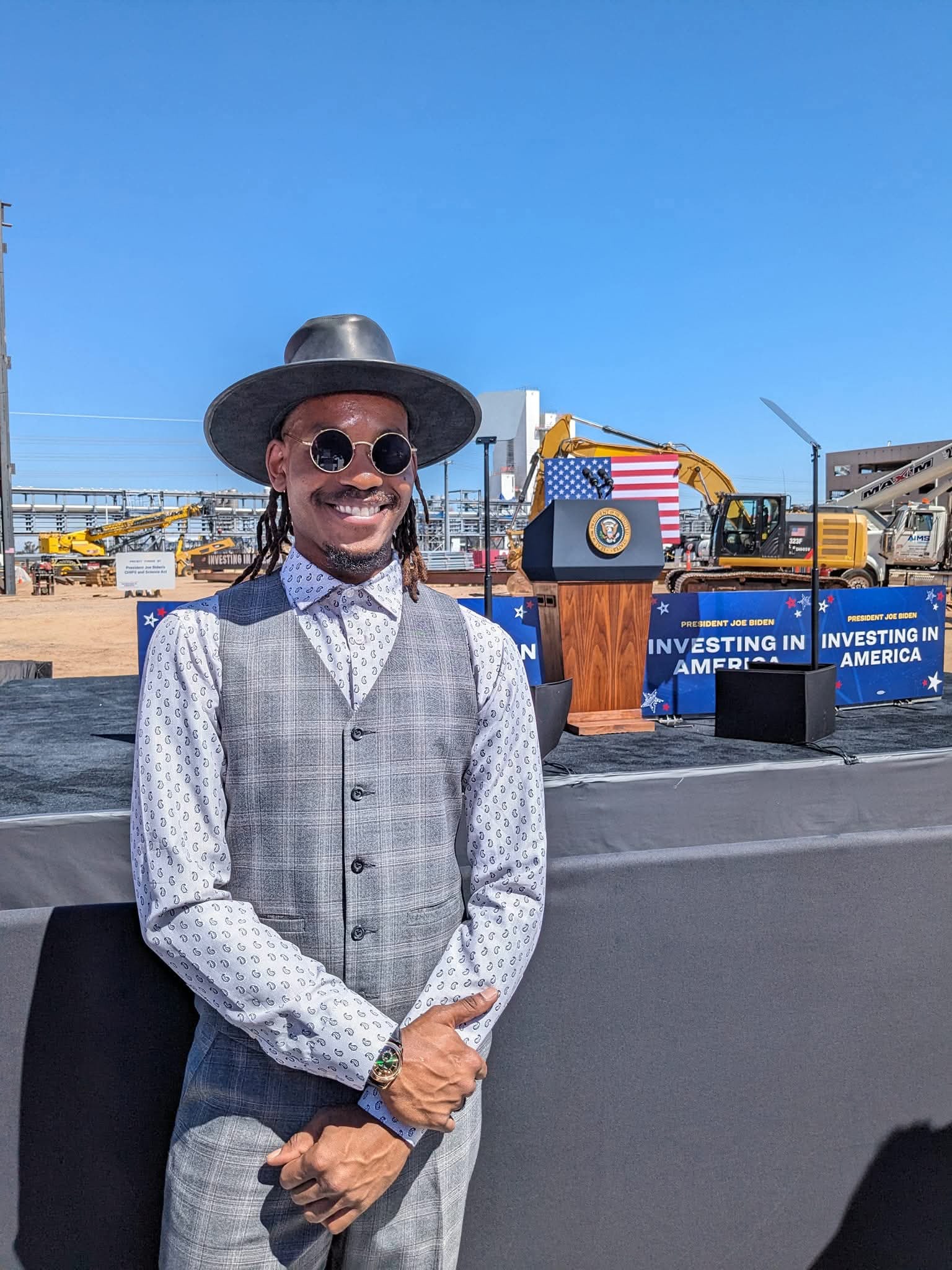 Rahmaan Phillip in a wide-brim hat and round sunglasses at a presidential event, the podium behind him.